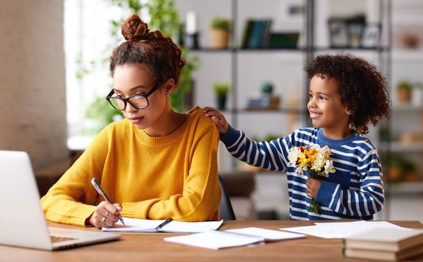 Surprise for mommy from son. Cute little mixed race boy with flower bouquet in hand going to congratulate mom on Mothers or Womens day while she working on laptop at home. Family celebration concept - Photo, Image