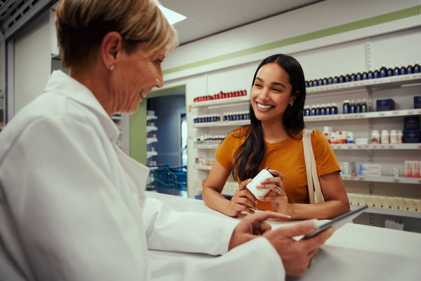 Happy senior female pharmacist showing screen of digital tablet to young customer holding bottle of supplements in pharmacy while in a conversation - Foto, imagen