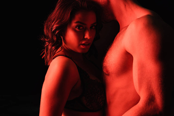 iluminación roja en la mujer sexy en lencería cerca del hombre muscular aislado en negro  - Foto, Imagen