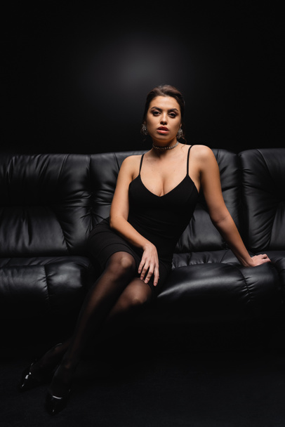 full length of seductive woman in slip dress sitting on leather sofa on black - Fotó, kép