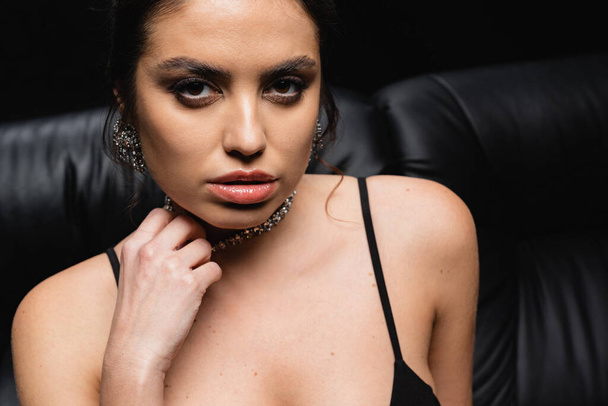 seductive woman adjusting necklace and looking at camera on black - Фото, изображение