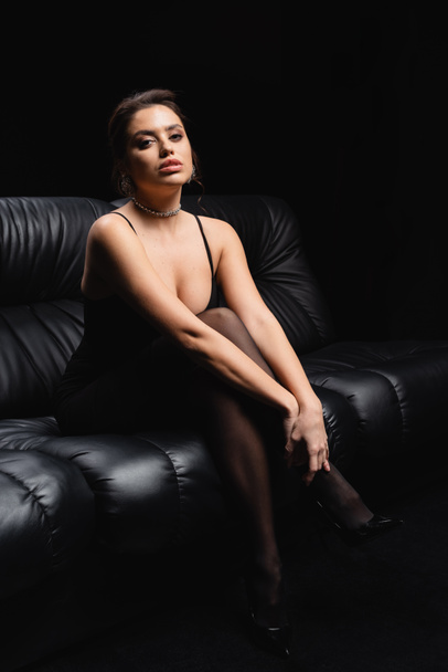 full length of seductive woman in slip dress and tights sitting on sofa on black - Fotografie, Obrázek