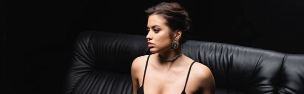 sexy woman in necklace and earrings sitting on sofa isolated on black, banner - Φωτογραφία, εικόνα