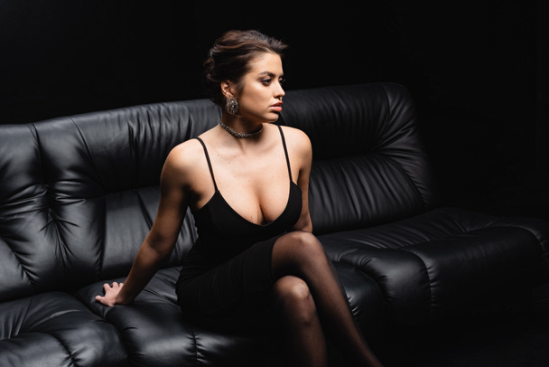 sexy woman in slip dress and tights sitting on leather sofa isolated on black - Foto, Imagen
