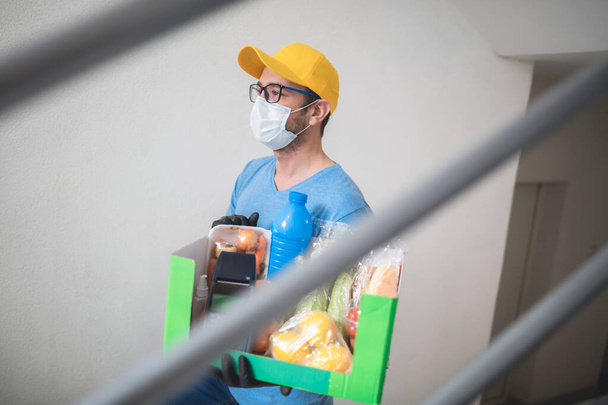 livreur sur l'escalier du bâtiment avec masque de protection tenant boîte avec épicerie et point de vente pour le paiement sans contact. - Photo, image