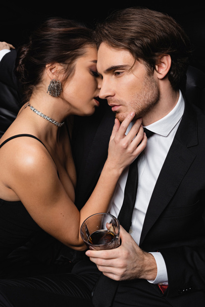 woman in slip dress seducing man in formal wear holding glass of whiskey on black  - Fotografie, Obrázek