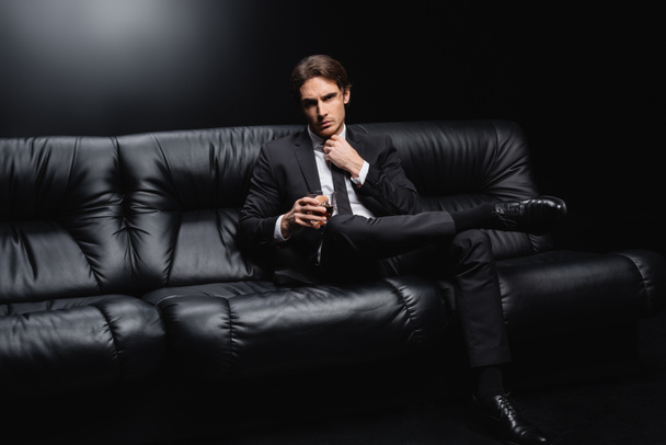 elegant man in suit adjusting tie and sitting on leather couch with glass of whiskey on black background  - Photo, Image