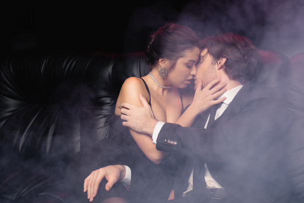 passionate woman in slip dress kissing man in suit on black with smoke - Photo, Image