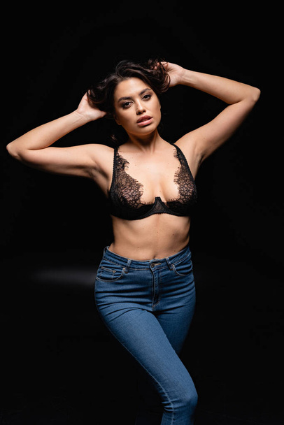 young brunette woman in lace bra and jeans looking at camera on black  - Photo, Image