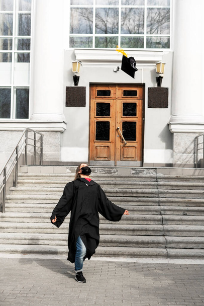 Siyahi mezuniyet elbisesi ve koruyucu yüz maskesi takan mutlu genç bir kadın üniversite ya da üniversite girişinin arka planında sarı püsküllü mezuniyet şapkası takıyor. Yeni, normal, gerçek insanlar. - Fotoğraf, Görsel