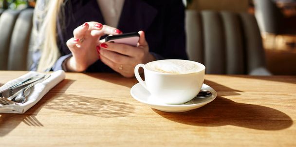 Woman typing text message on smart phone in a cafe. Young woman sitting at a table with a coffee using mobile phone. Coffee break. Cup of coffee on the table. Blurred image, selective focus - Фото, зображення