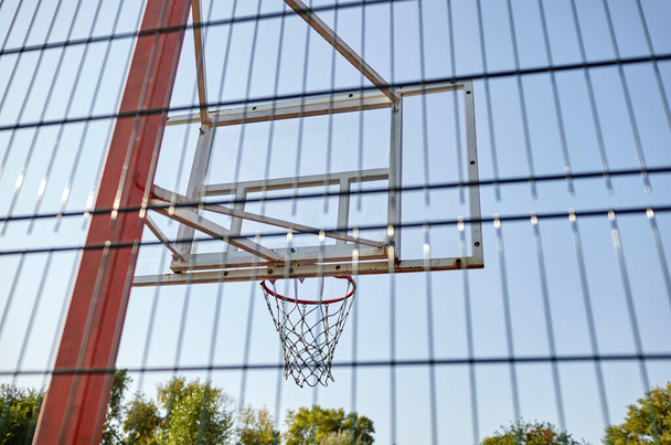 Primer plano aro de baloncesto contra el cielo azul en el parque de la ciudad. Cancha de baloncesto detrás de la valla. Enfoque selectivo, fondo borroso - Foto, imagen