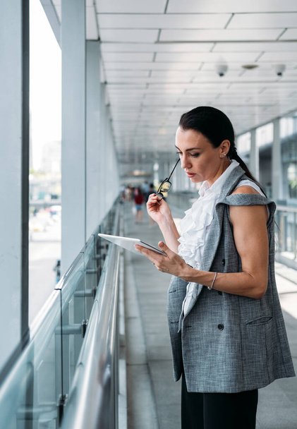 Ділова жінка використовує планшет на сучасному балконі. Портрет красивої бізнес-леді, що тримає окуляри і дивиться на планшет
 - Фото, зображення