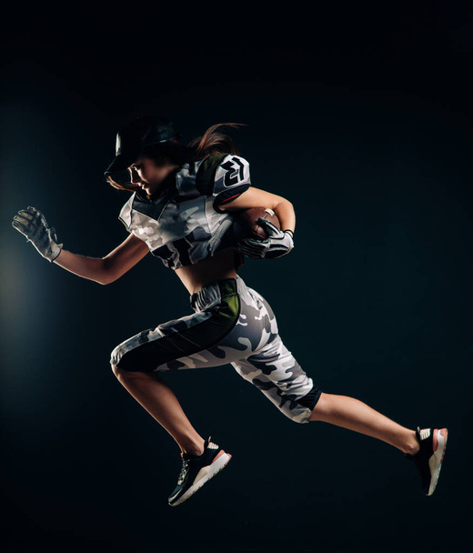 Mooie sexy atletische vrouw loopt op zwarte achtergrond in Amerikaans voetbal uniform en pet met bal in haar handen. - Foto, afbeelding