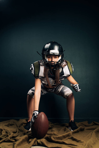 Hermosa mujer atlética sexy posa sobre fondo negro en uniforme de fútbol americano y casco con bola en la mano. - Foto, Imagen