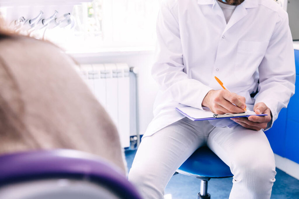 tandarts zit in stoel en registreert de reacties van de patiënt. tandheelkundige kliniek. geschiedenis te nemen. - Foto, afbeelding