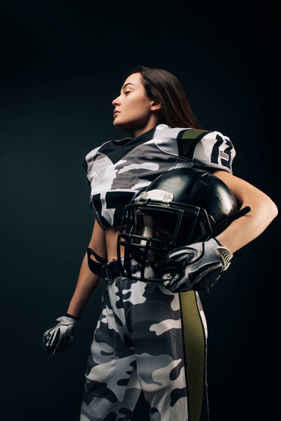 Hermosa mujer atlética sexy posa sobre fondo negro en uniforme de fútbol americano con casco en la mano. - Foto, Imagen