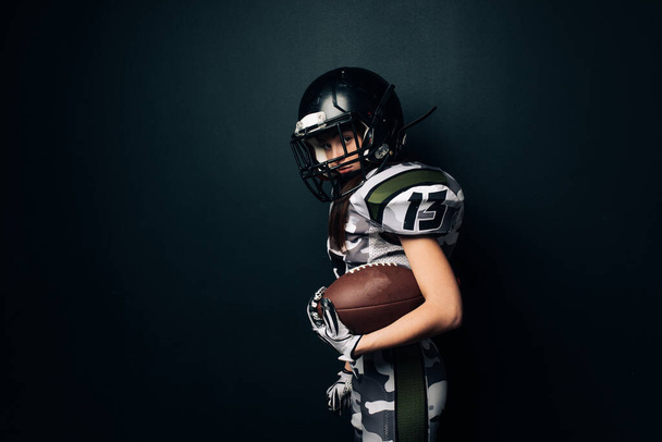 Hermosa mujer atlética sexy posa sobre fondo negro en uniforme de fútbol americano y casco con bola en la mano. Vista lateral. - Foto, imagen