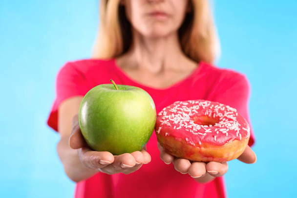 Woman choosing between doughnut and fresh apple on light blue background, closeup - Foto, Imagen