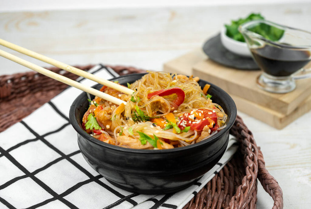 Asian salad with rice noodles, vegetables, mushrooms, chicken and soy sauce. Funchose with white transparent noodles in black plate - Photo, Image