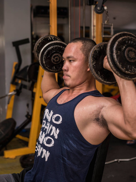 A athletic guy leans on an incline bench while doing seated shoulder dumbbell presses. - Fotó, kép
