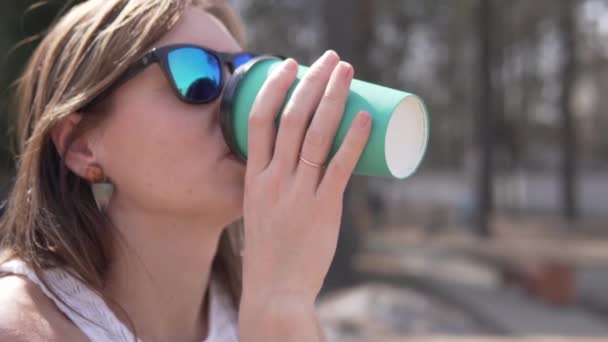 Een vrouw met een zonnebril drinkt in het voorjaar koffie uit een plastic glas op straat - Video