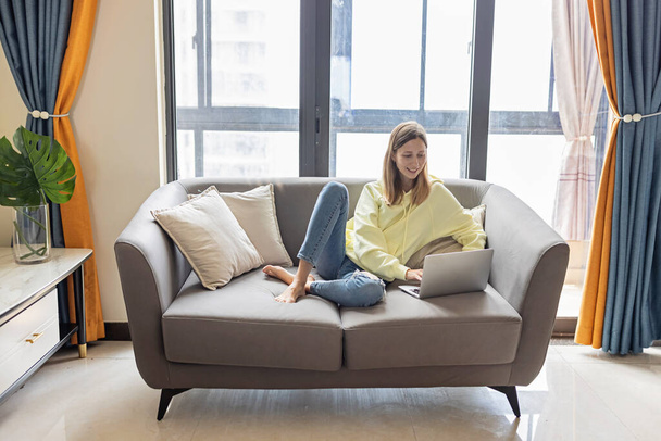 Young beautiful woman wearing yellow hoodie and resting on gray textile sofa at home. Attractive slim female in domestic situation, staying home, relaxing on couch in her apartment. Background, copy - Photo, image