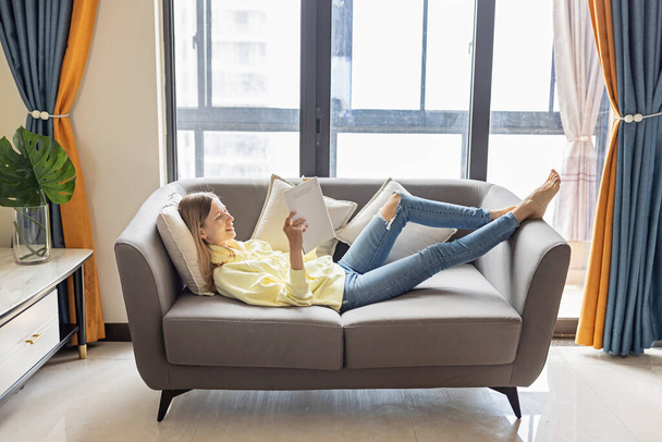 Young beautiful woman wearing yellow hoodie and resting on gray textile sofa at home. Attractive slim female in domestic situation, staying home, relaxing on couch in her apartment. Background, copy - Photo, Image