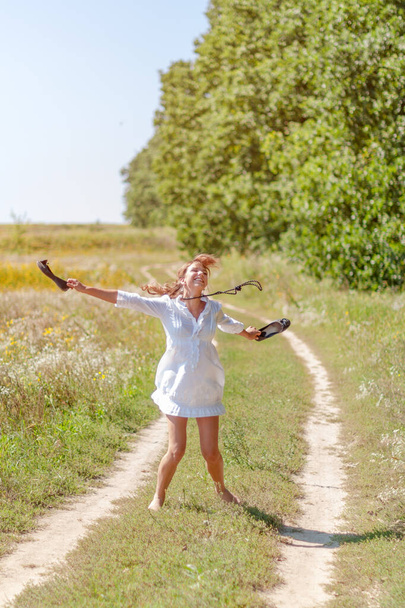 Joven hermosa mujer corriendo en la hierba descalza y sosteniendo zapatos en las manos. - Foto, Imagen
