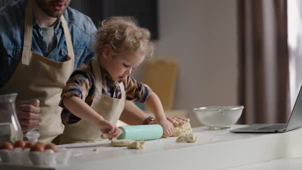χαριτωμένο νήπιο με αστεία μπούκλες παίζει στην κουζίνα, τροχαίο έξω ζύμη, master-class από τον πατέρα, τον μπαμπά και τον γιο - Πλάνα, βίντεο