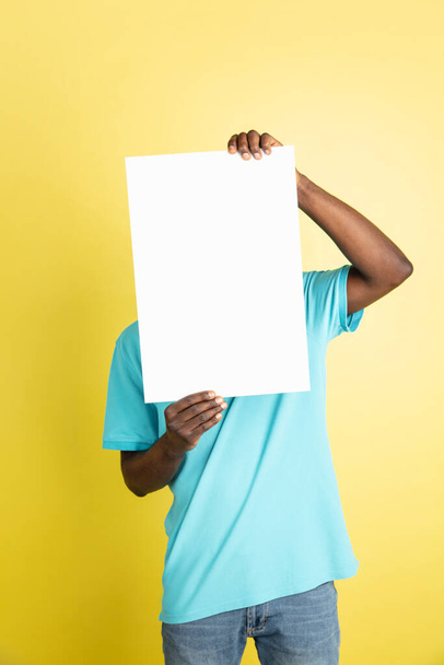 Young African man with blank sheet of paper isolated over yellow studio background. - 写真・画像