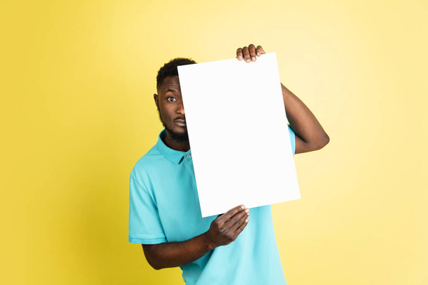 Young African man with blank sheet of paper isolated over yellow studio background. - 写真・画像