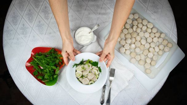 Hands of a woman hold out a bowl of cooked homemade dumplings, decorated with green leaves of dill and parsley against background of white round table with raw dumplings on cutting board. Flat lay. - Photo, Image