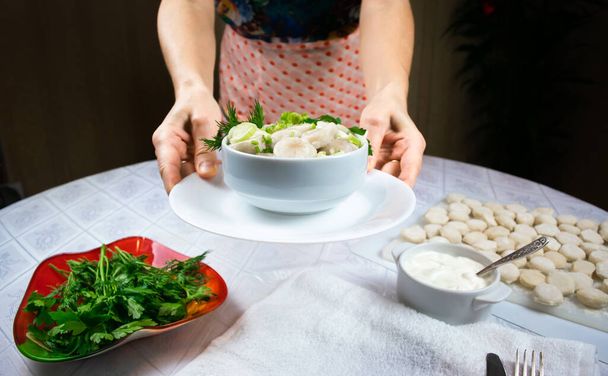 Hands of a woman in an apron hold out a bowl of cooked homemade dumplings, decorated with green leaves of dill and parsley against background of a round table with a white tablecloth. Selective focus. - Photo, Image