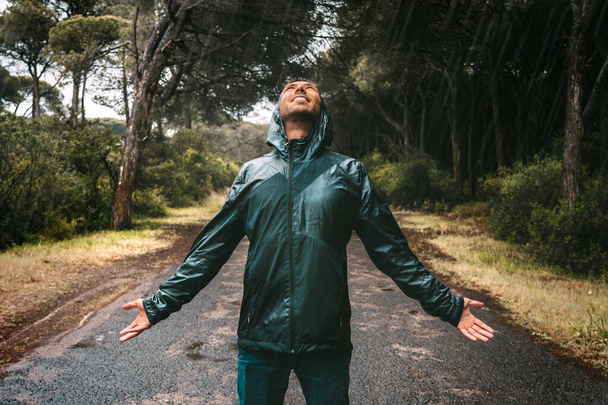 Young wet smiling man in drenched jacket with a hood enjoying rain. Man in raincoat stood in heavy rain. Bad autumn wether. - Photo, Image