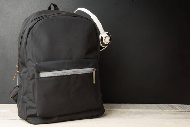 рюкзак с наушниками на фоне черной стены, на белом деревянном столе - Фото, изображение