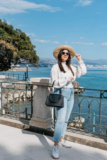 Attractive young woman in white blouse and blue jeans standing on terrace on sea shore. Young tourist standing on seafront in Opatija, Croatia. - Foto, imagen