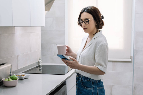 donna che beve caffè in cucina mentre usa il suo smartphone - Foto, immagini