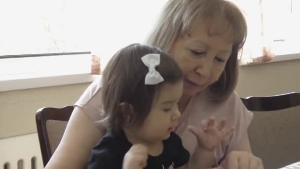 Een oudere vrouw met een klein kind zit aan tafel. De grootmoeder toont het kind foto 's en leert. - Video