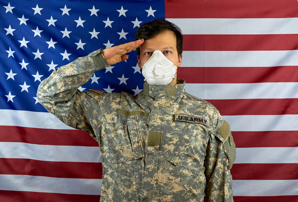 A soldier in a respirator salutes. American soldier salutes, wearing a protective mask against COVID-19 on the background of the American flag. - Photo, Image