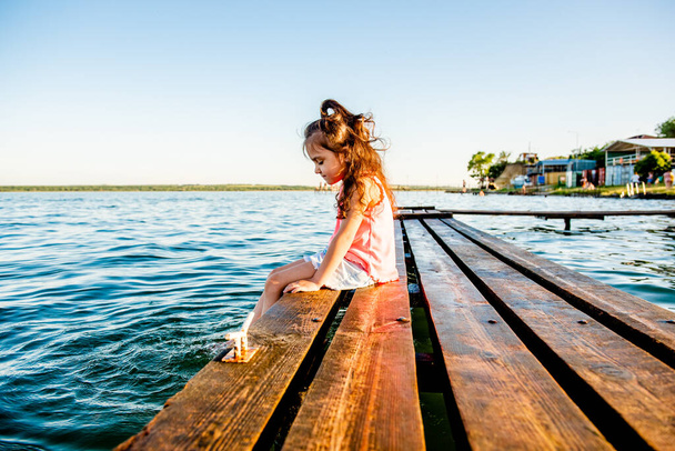 Cute little girl on the beach. The kid is resting by the sea. Summer vacation. Girl in the summer. A beautiful little girl sits on the pier by the water. girl lying on a wooden bench on the beach.  - Photo, Image