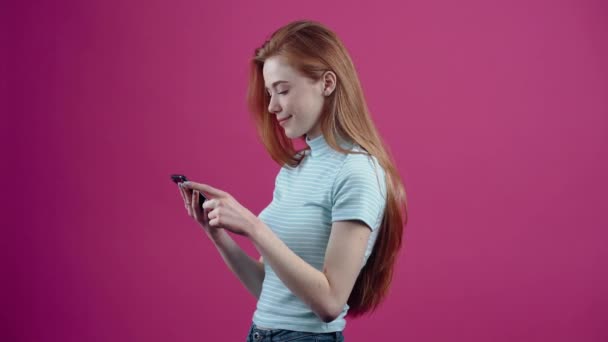In profile, a young woman shocked by the phone in her hand follows the news and smiles broadly, approving with her thumb up, in a blue casual T-shirt, isolated on a pink background. The concept of - 映像、動画