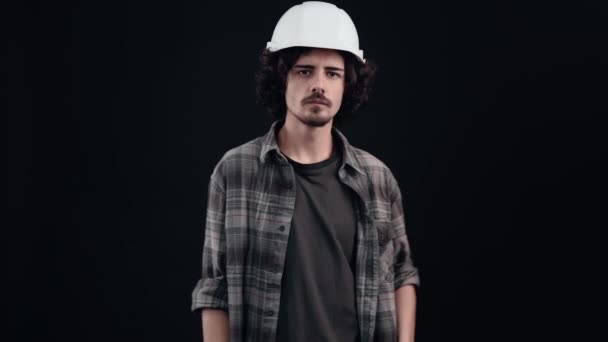 Young worker with his finger on his lips says to shut up, quietly. He wears a construction helmet and a closed shirt on a black background in the studio, Industrial concept, employees - Footage, Video