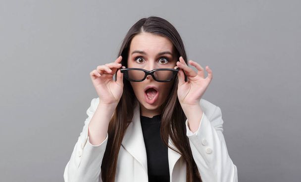 white funny young woman in a white jacket with glasses is surprised on a gray background - Φωτογραφία, εικόνα