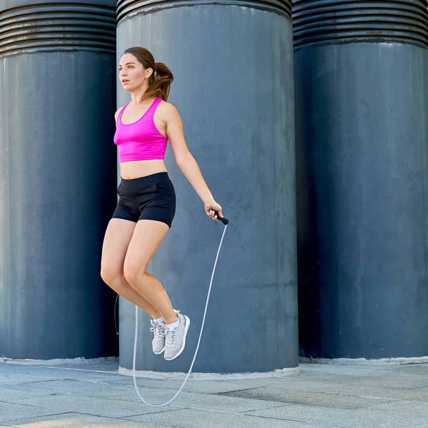 A focused, purposeful young pretty girl in sporty pink top and shorts, jumping rope in the summer, burns calories, loses weight, keeps herself in shape. Sports concept in quarantine, physical health. - Photo, Image