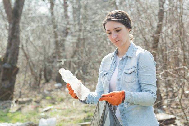 Female volunteer looks anxiously at the camera, and holds a bag with collected plastic, cleaning the forest and park from garbage - 写真・画像