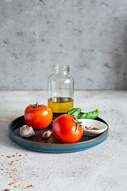 Fresh ripe vegetables (garlic,tomatoes) with basil leaves, spices and olive oil on a plate on a gray background. Healthy eating concept, raw food. Go vegan. A set of ingredients for preparing a salad. - Foto, imagen