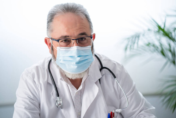 Portrait of senior bearded doctor in white coat and face mask looking at camera in his office. - 写真・画像