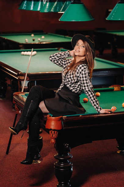 A girl in a hat in a billiard club sits on a billiard table.Playing pool - Φωτογραφία, εικόνα