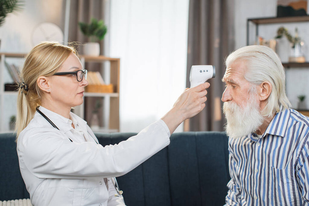 Confident 40-aged blond woman doctor checking body temperature of mature bearded man patient using non-contact infrared forehead thermometer during home visit - Zdjęcie, obraz